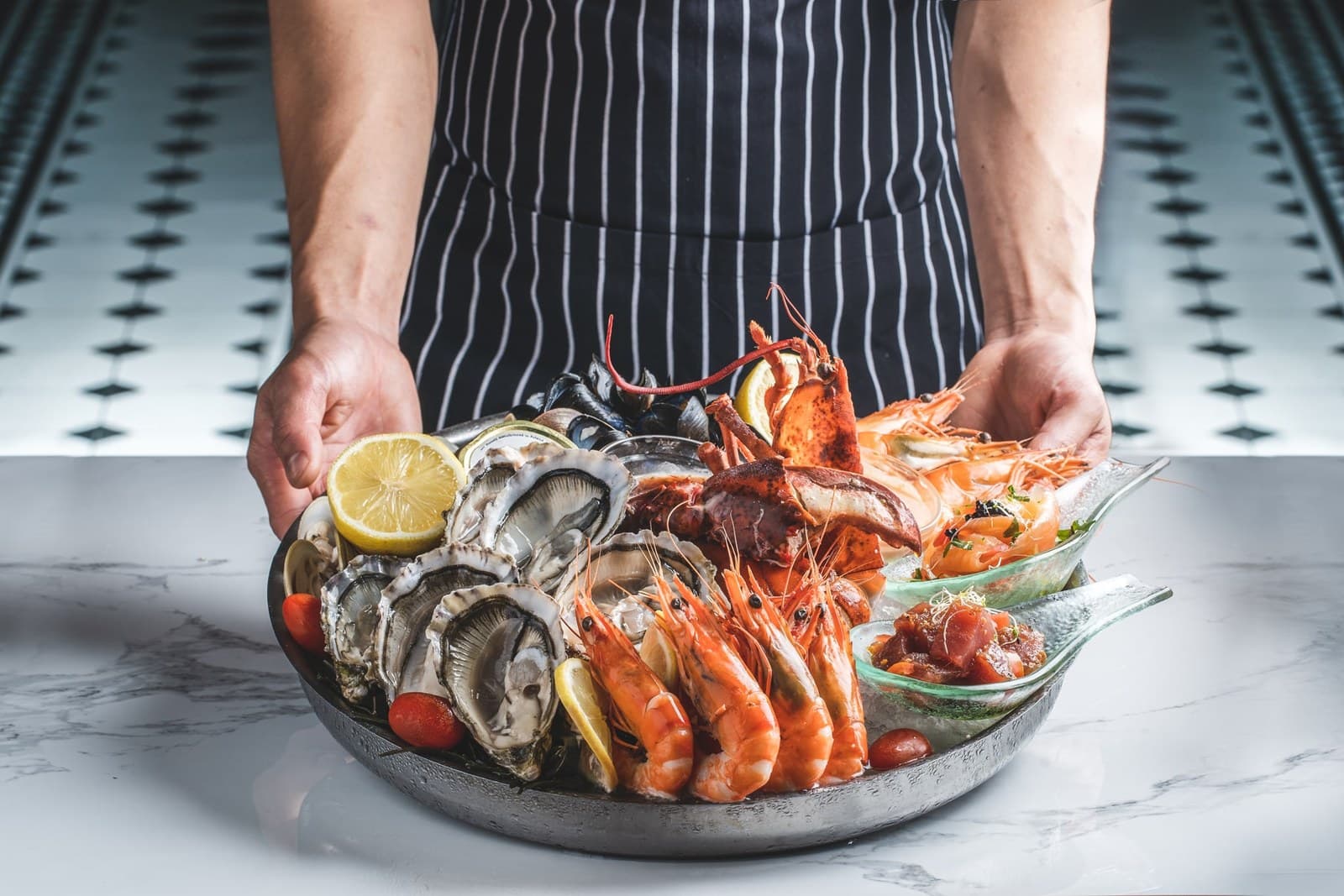 A chef wearing a navy-and-white striped apron holds a large round black ceramic platter brimming with chilled seafood: half-shell oysters with lemon, jumbo prawns, mussels, lobster claw, scallops, and salmon sashimi-style strips. White marble surface in foreground with black-and-white patterned tile floor visible behind.