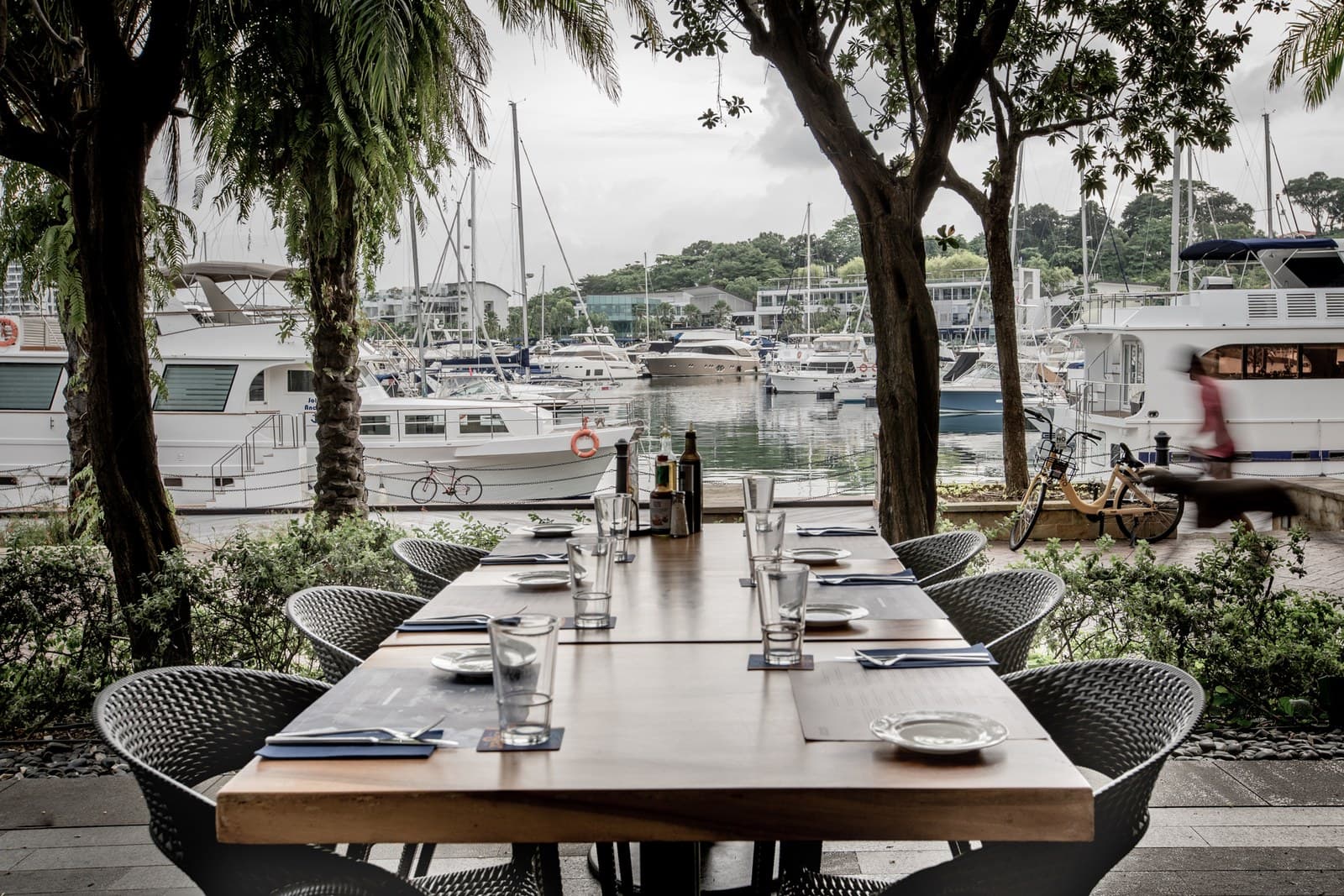 Alfresco dining setup at Greenwood Fish Market Quayside Isle (Sentosa) — long wooden table set for 6+ with grey wireframe chairs, placemats, water glasses, and a salt-and-pepper caddy. Background shows the Quayside Isle marina with white yachts moored at the boardwalk and Sentosa Cove residential buildings beyond. Tall palm trees frame both sides of the view.