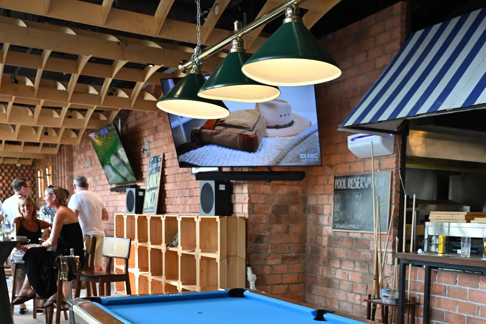 Interior of Greenwood Sports Bar at Quayside Isle showing a regulation pool table with bright blue felt under three green-shaded billiard lamps. The ceiling has exposed wooden arched beams, brick walls run along the right side with a striped white-and-blue awning, and multiple flat-screen TVs are mounted on the back wall. Casual diners sit at high stools in the background.