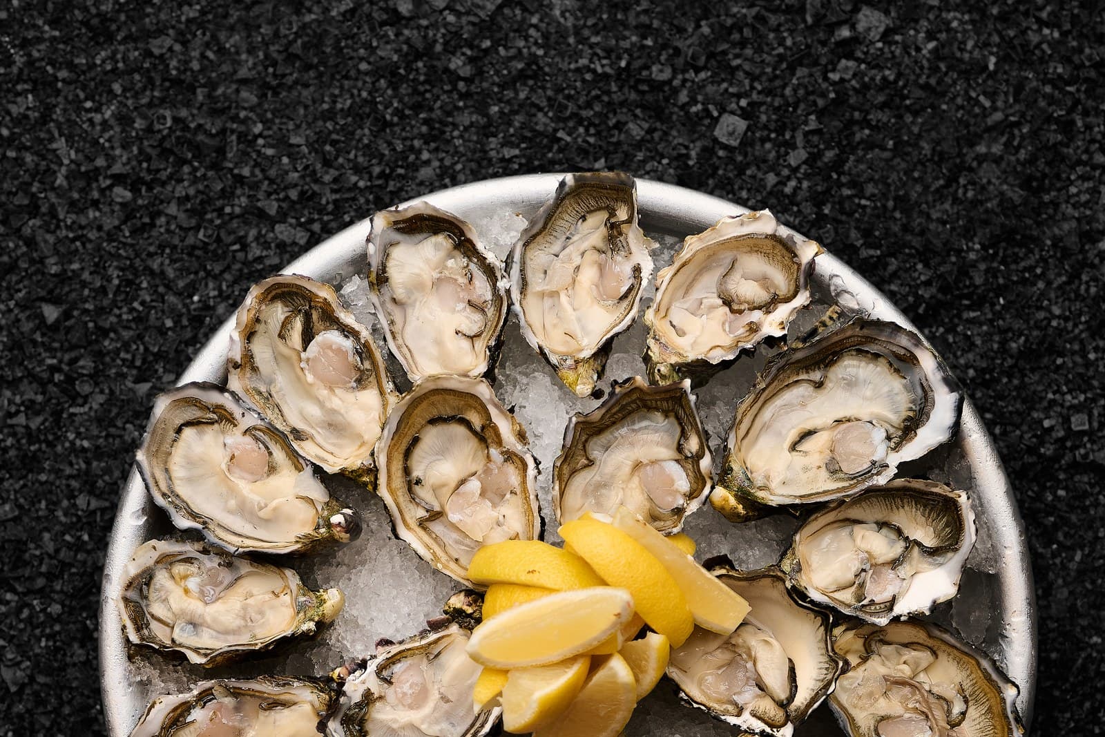 Overhead view of a metal platter filled with freshly-shucked Tasmanian oysters arranged in a circular pattern on crushed ice. A small mound of lemon wedges sits in the centre. The dark textured background suggests a marble or stone serving surface.