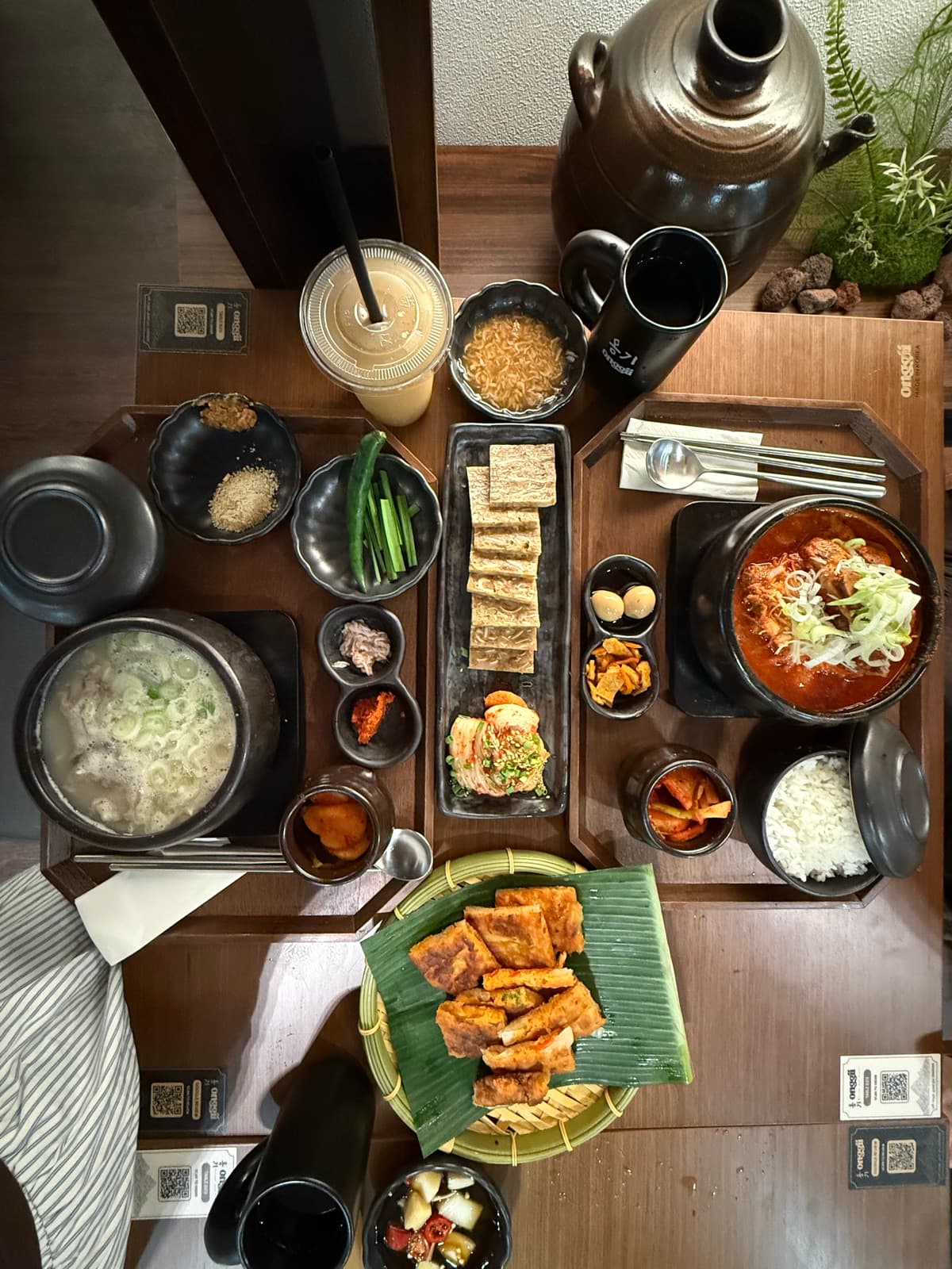 俯拍 Onggii 餐桌:牛骨汤、辣炖锅、香蕉叶煎饼、小菜、onggi 陶碗白饭与柚子饮。