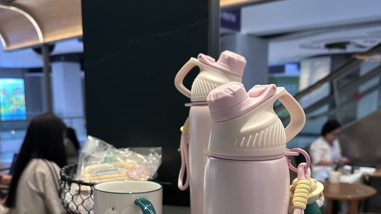 Wide in-store view: the new sakura-pink Starbucks bottle with a 3D Bearista bear charm clipped to the cap (right), paired with two ceramic mugs featuring scattered cherry/berry patterns. The Starbucks storefront is visible in the background.