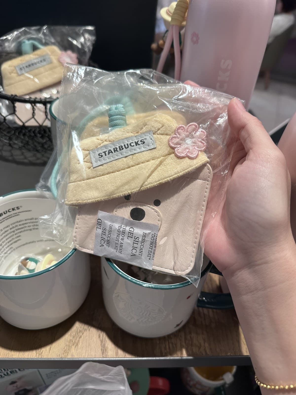 Close-up of a hand holding a small fabric pouch shaped like a Bearista bear with a knitted hat. The pouch has a small pink flower decoration and the Starbucks logo tag. A small bear face is visible on the pouch's front, with a desiccant gel pack tucked inside the clear packaging.