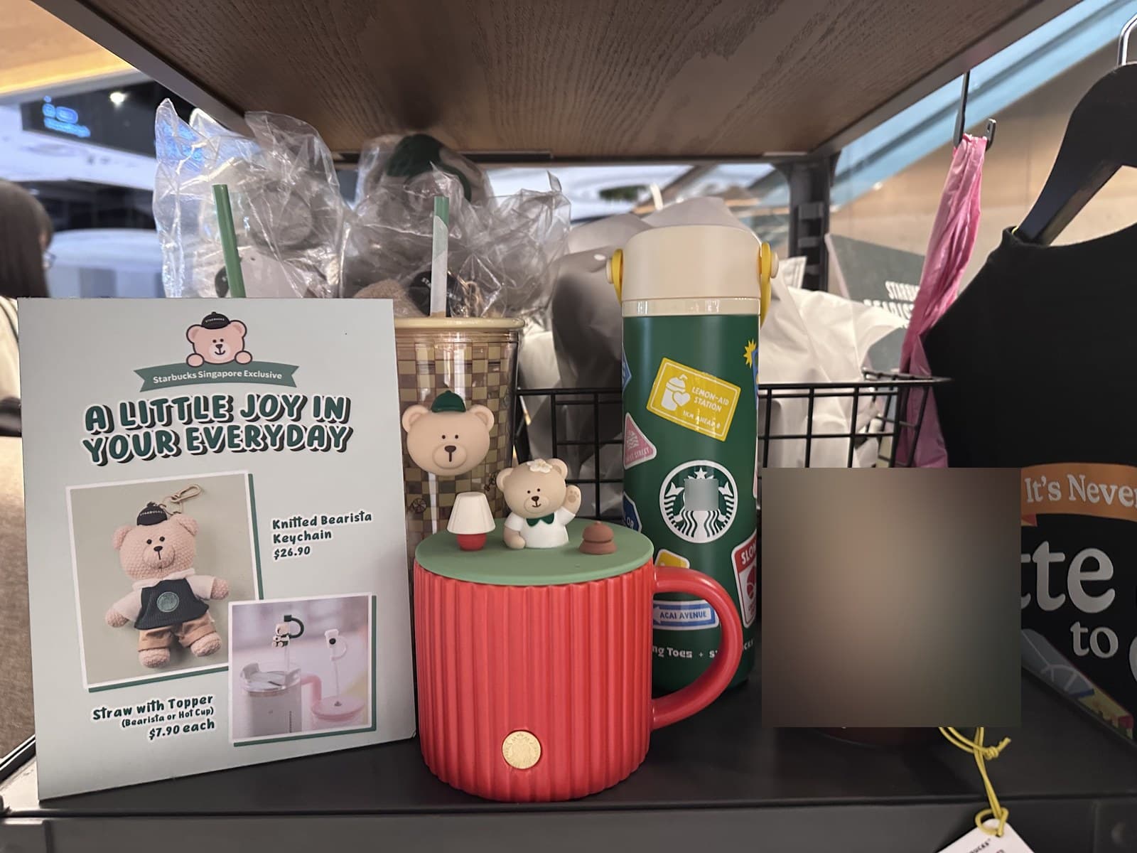 In-store display showing the Starbucks "A Little Joy in Your Everyday" Singapore Exclusive campaign. Left side: the Knitted Bearista Keychain priced at S$26.90 with a separate display of the Straw with Topper (Bearista or Hat Cup) priced at S$7.90 each. Centre: a small ceramic mug with two 3D bear figurines (one in a hat, one in green apron) on top of the lid. Right side: a green stainless tumbler with various campaign stickers.