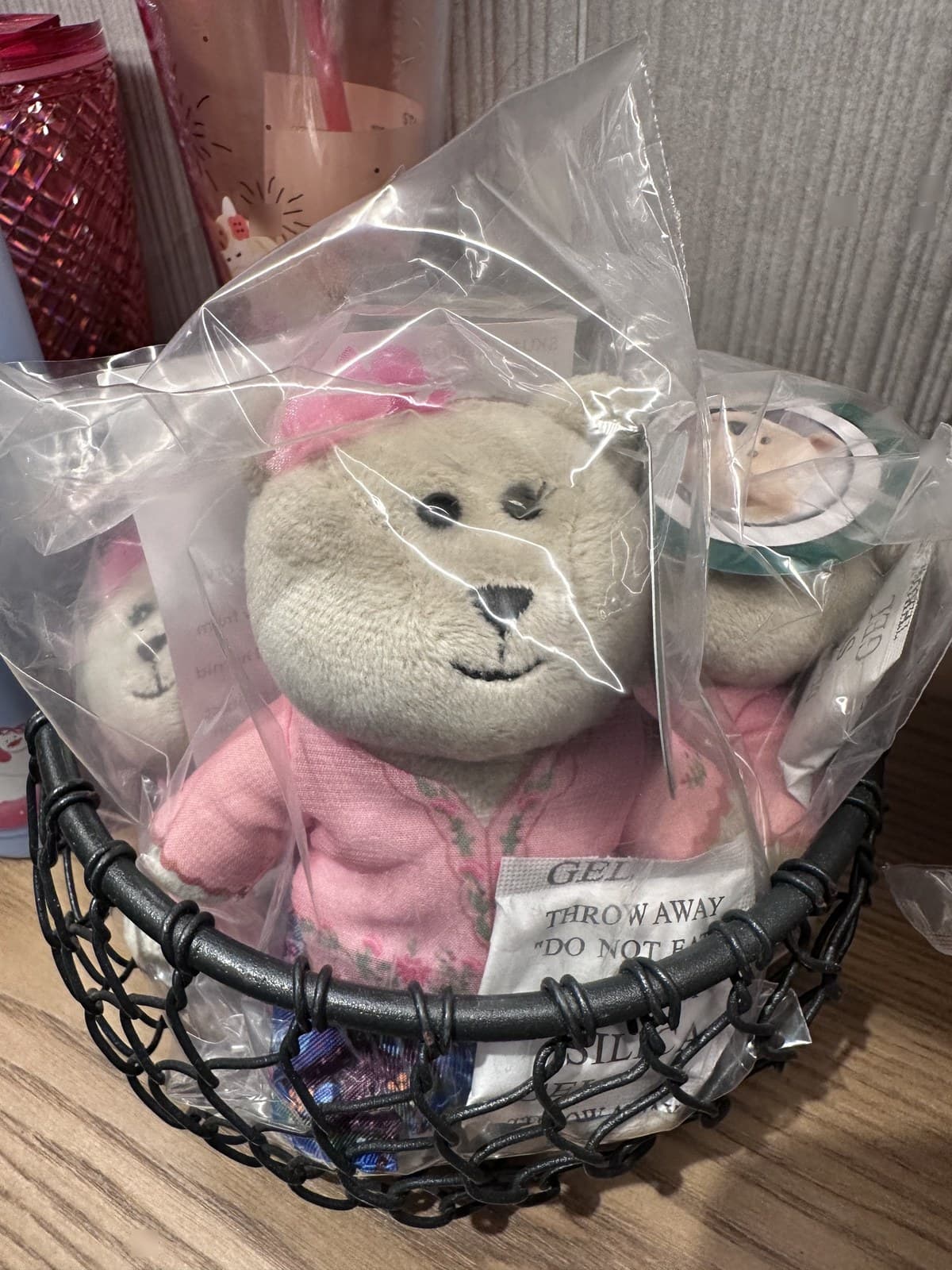 Close-up of a plush Bearista teddy bear wearing pink pyjamas and a Starbucks barista uniform. The plush is individually packaged in a clear plastic bag and displayed in a black wire basket with several other identical packaged bears visible.