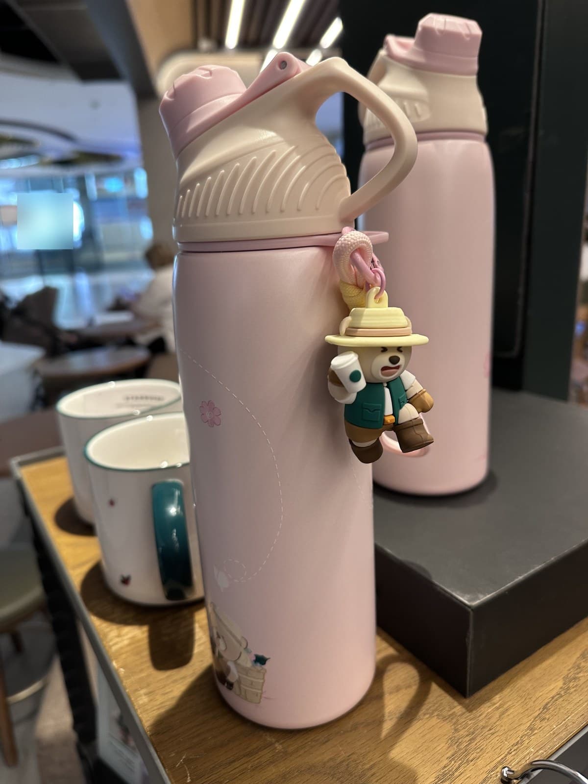 Close-up of the sakura-pink Starbucks bottle with a 3D Bearista bear charm clipped to the cap. The bear character holds a small bucket and wears a hat (adventure/camping themed). Two ceramic Starbucks Siren-logo mugs are visible in the foreground, with a second pink bottle in the background.