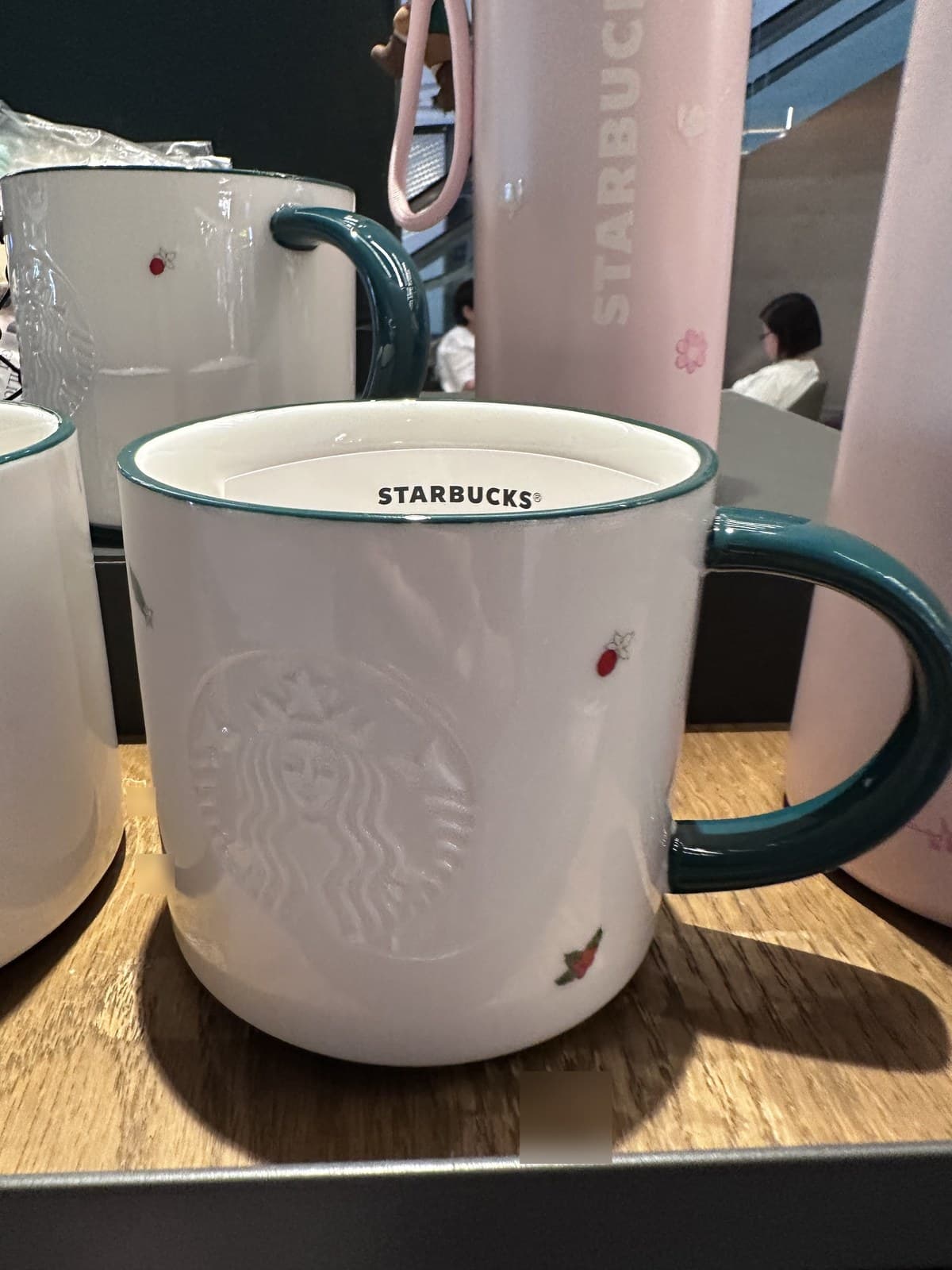 Close-up of a white Starbucks ceramic mug with a teal/aqua handle. The mug features an embossed Starbucks Siren logo and small scattered cherry/berry print accents.