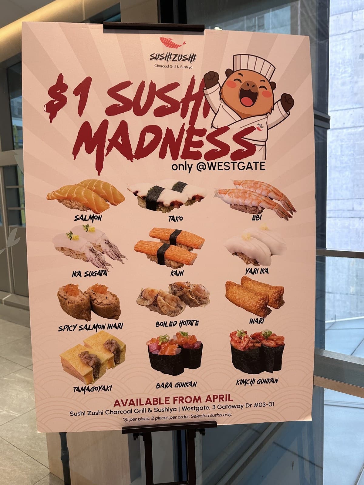 Standee photo of the $1 SUSHI MADNESS poster. Lists 12 sushi items at $1 each: Salmon, Tako, Ebi, Ika Sugata, Kani, Yari Ika, Spicy Salmon Inari, Boiled Hotate, Inari, Tamagoyaki, Bara Gunkan, Kimchi Gunkan. Available from April. Footnote: $1 per piece, 2 pieces per order, selected sushis only. Address: Sushi Zushi Charcoal Grill & Sushiya, Westgate, 3 Gateway Dr #03-01.
