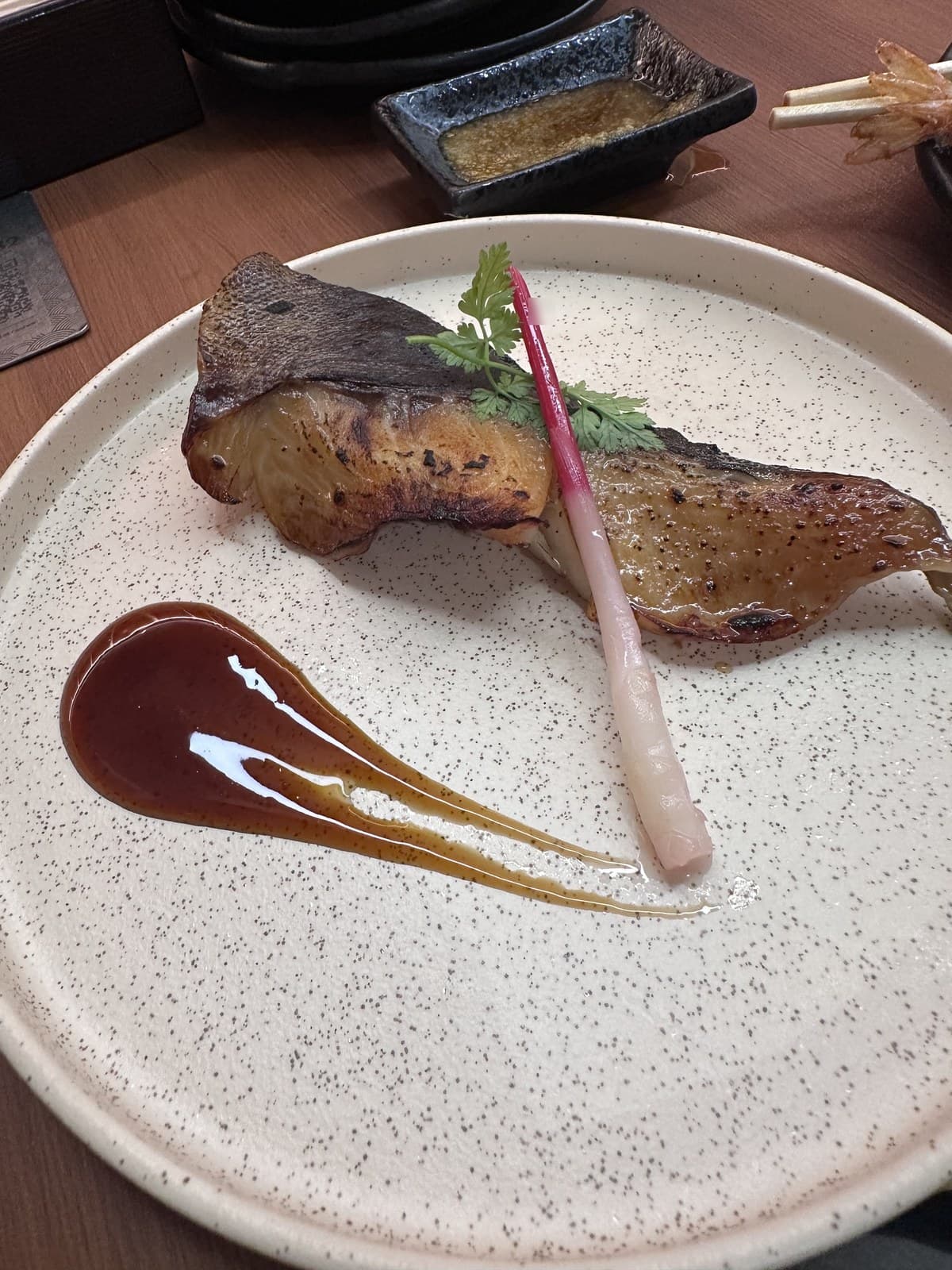 A small grilled fish dish on a textured beige plate — looks like a small portion of grilled cod or similar white fish, served with a vibrant pink pickled ginger garnish + green herb sprig. A pool of dark dipping sauce sits beside it. Small ceramic dish with more sauce in the background, plus the edge of bamboo skewers.