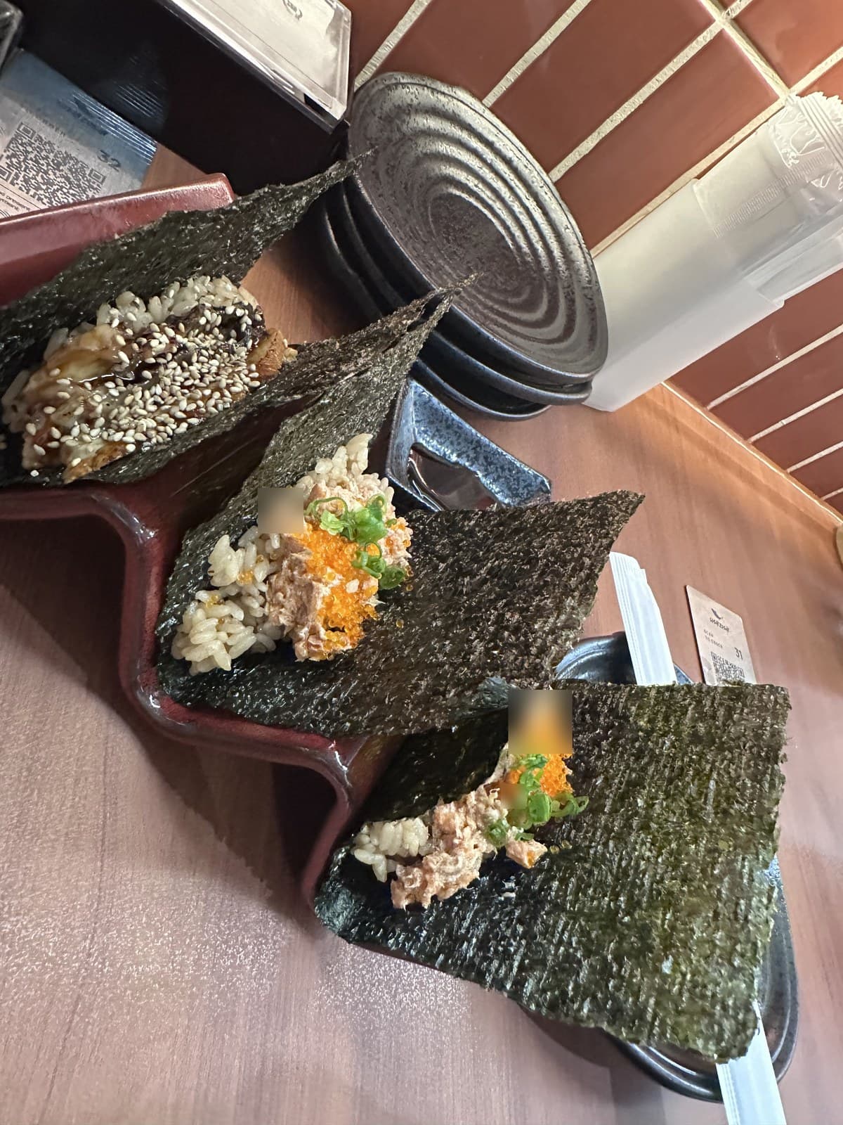 Close-up of three hand-rolled sushi (temaki) on a wooden tray. Each handroll is wrapped in dark nori and filled with sushi rice + various toppings (one with sesame seeds + tuna, one with mixed seafood + tobiko on a salad-style base). Stacks of black ceramic plates are visible in the background.