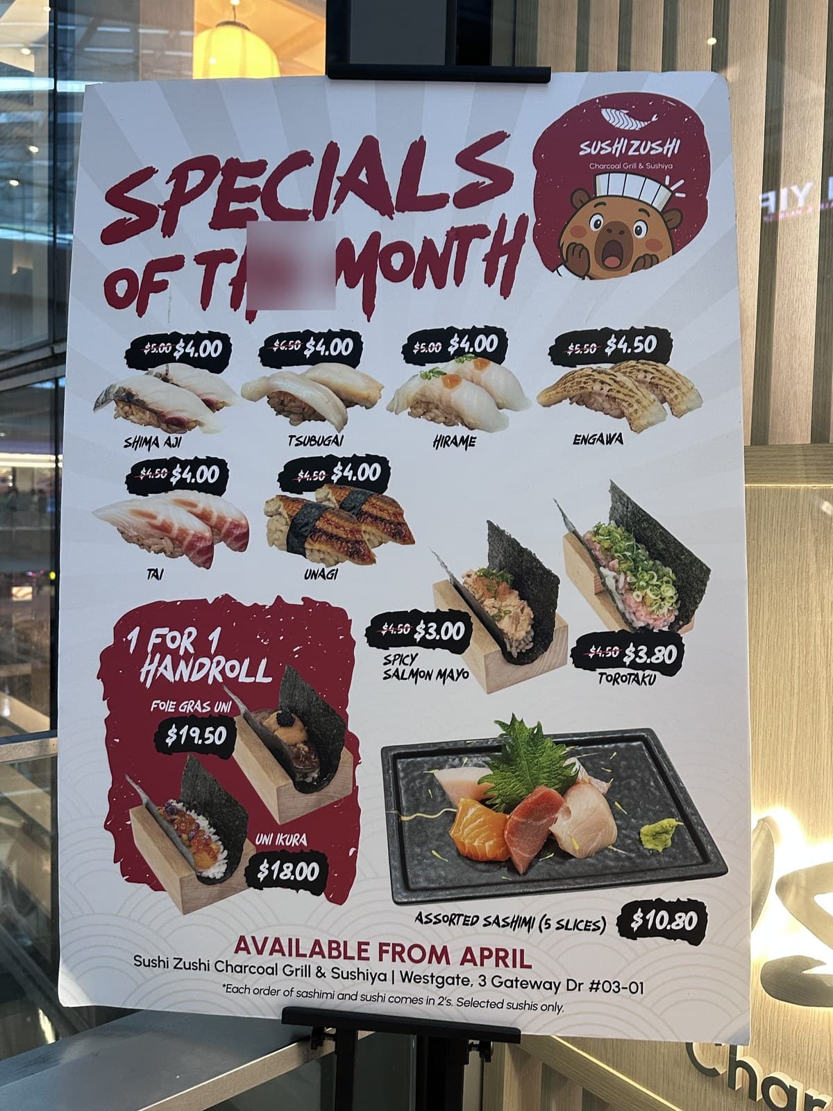 Standee photo of Specials of the Month poster. Six premium nigiri reduced: Shima Aji $5.00→$4.00, Tsubugai $5.00→$4.00, Hirame $5.00→$4.00, Engawa $5.50→$4.50, Tai $4.50→$4.00, Unagi $5.50→$4.00. Plus 1-for-1 handroll on Foie Gras Uni ($19.50) and Uni Ikura ($18). Spicy Salmon Mayo handroll $4.50→$3.00, Torodaku $4.50→$3.80. Assorted Sashimi (6 slices) $10.80. Available from April. Same Westgate address.