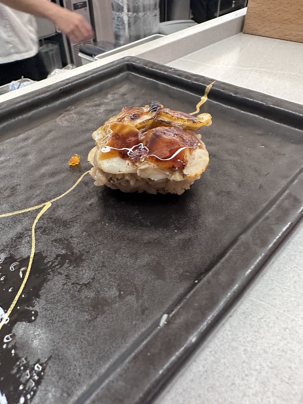 黑陶瓷盘上单个鰻握寿司近照。鰻有明显烤痕+ 亮甜酱,放在小饭团上。盘表黄酱拖痕。厨师围裙在上背景部分可见。