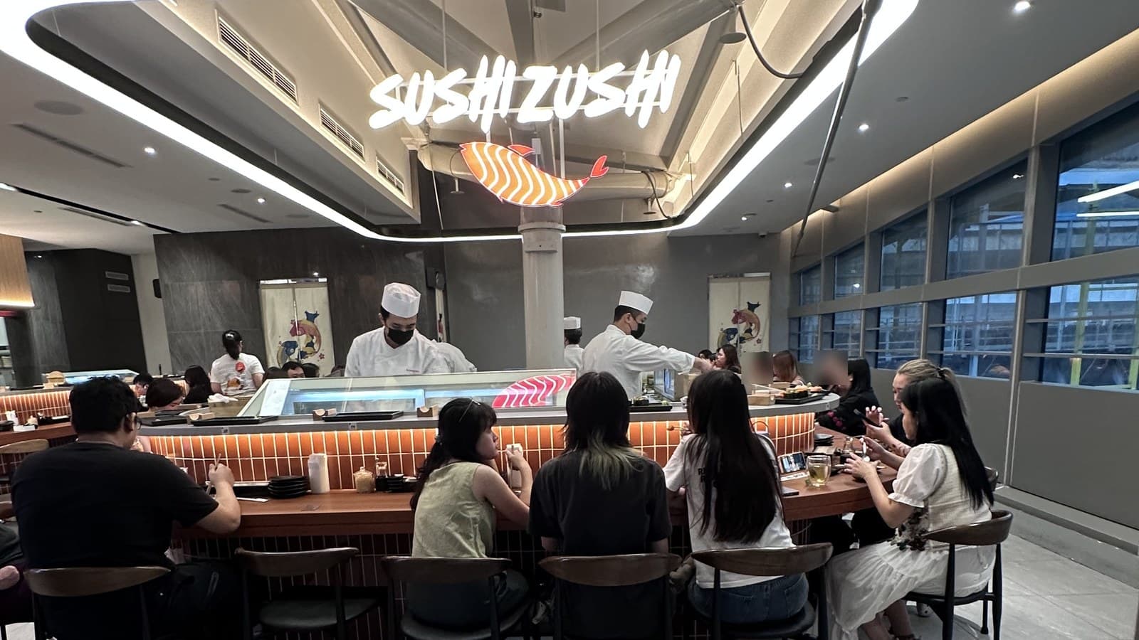 Sushi Zushi @ Westgate 广角内部。明亮三角形 SUSHIZUSHI 标志挂在寿司吧上方,2 位白围裙+ 帽厨师在柜台工作。数位顾客背对镜头坐在柜台(脸已模糊保护隐私)。商场走廊与 Westgate 玻璃幕墙在右。
