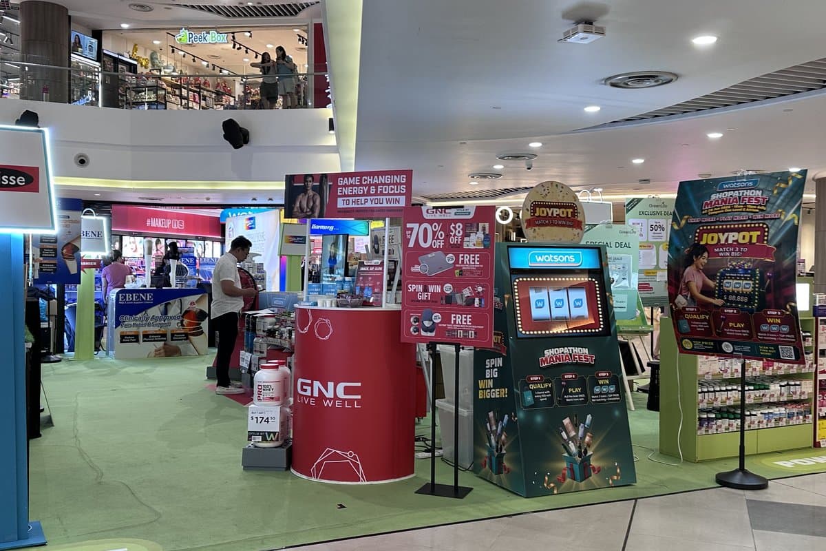 The GNC booth with 70%-off and spin-to-win signage next to the lit, arcade-style digital JoyPot game machine and Shopathon Mania Fest standees.