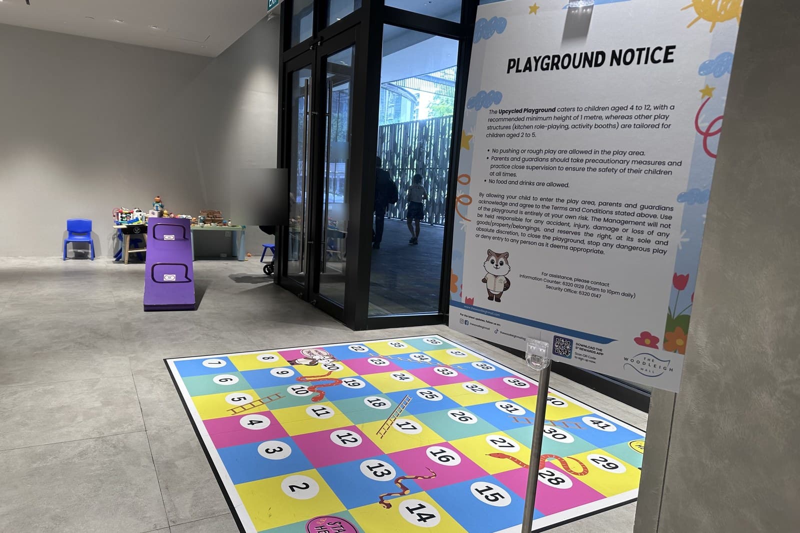 游乐场入口含 Playground Notice 标志(规则+ 年龄指引)与门槛上的蛇梯棋地贴。透过玻璃门可看到通往商场其余的走廊。