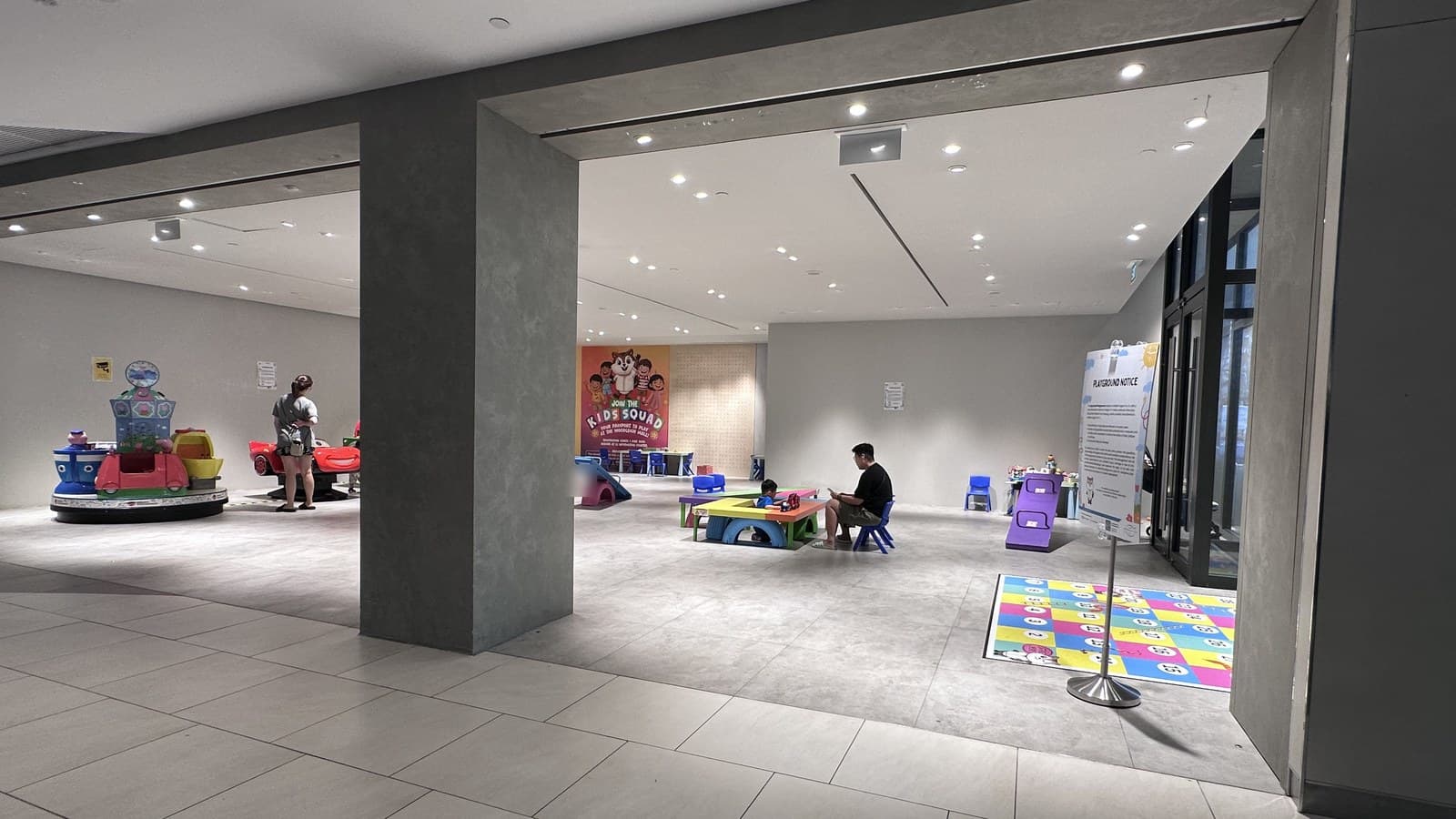 Woodleigh Mall「The Upcycled Playground」的广角全景 — 旋转平台迷你乘骑车(左)、KIDS SQUAD 吉祥物墙(中)、有小孩玩耍的厨房角色扮演桌(中右)、蛇梯棋地贴(右)。入口可见 Playground Notice 标志。