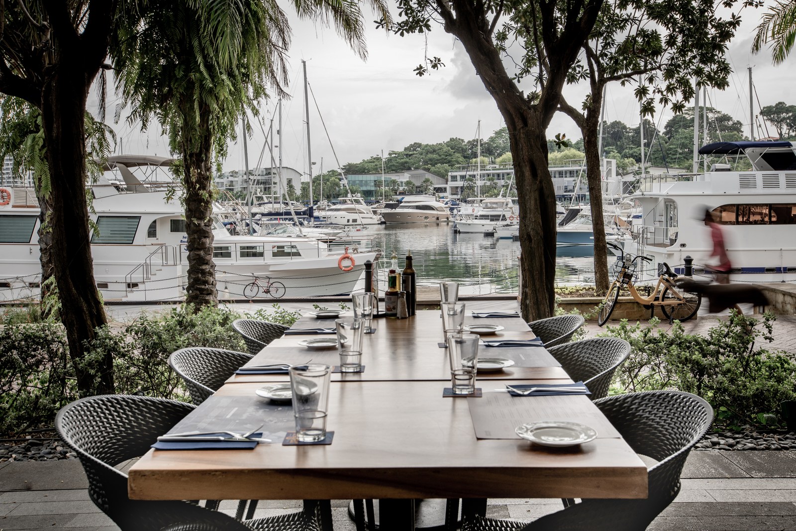 Alfresco dining setup at Greenwood Fish Market Quayside Isle (Sentosa) — long wooden table set for 6+ with grey wireframe chairs, placemats, water glasses, and a salt-and-pepper caddy. Background shows the Quayside Isle marina with white yachts moored at the boardwalk and Sentosa Cove residential buildings beyond. Tall palm trees frame both sides of the view.