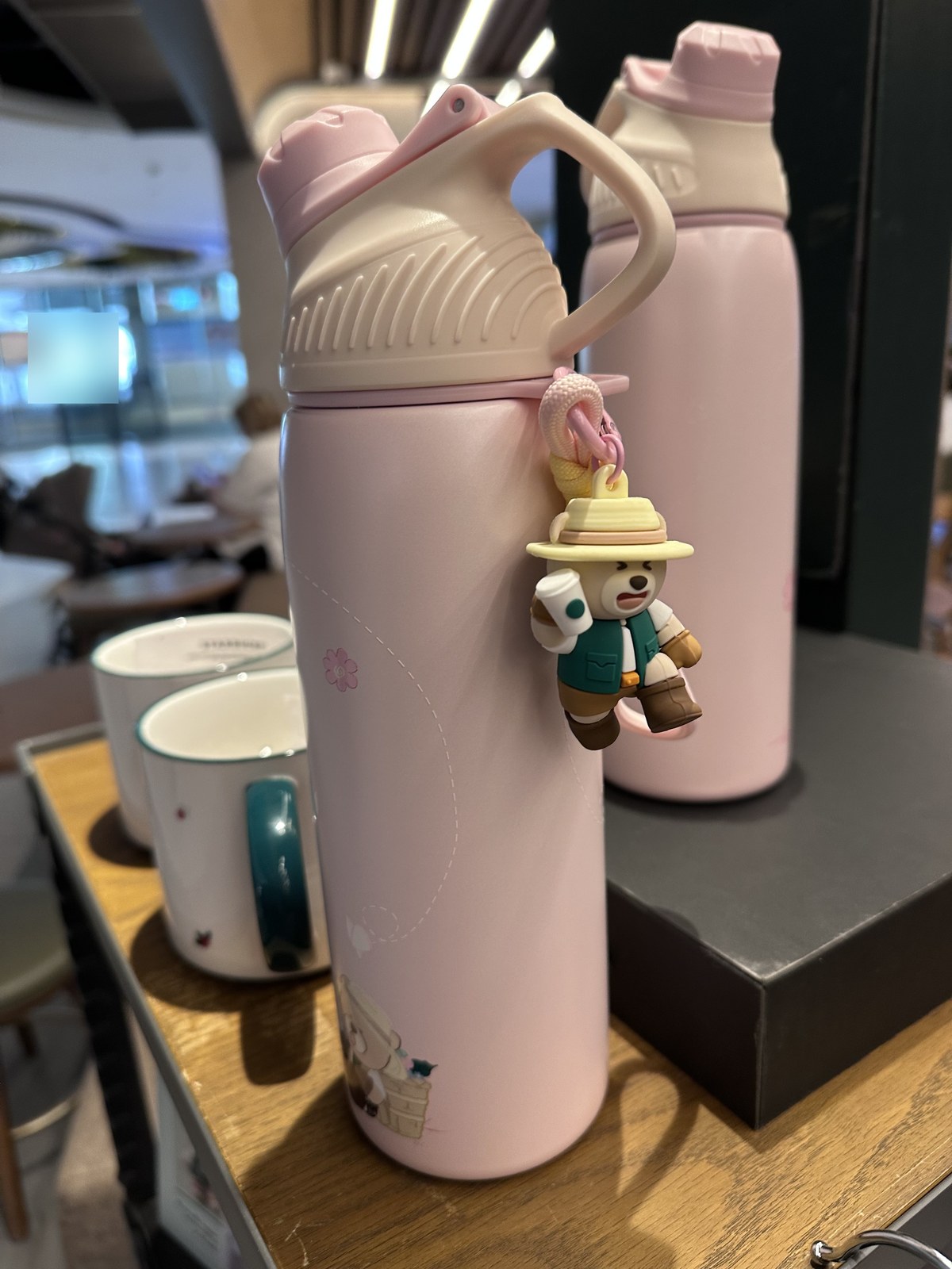Close-up of the sakura-pink Starbucks bottle with a 3D Bearista bear charm clipped to the cap. The bear character holds a small bucket and wears a hat (adventure/camping themed). Two ceramic Starbucks Siren-logo mugs are visible in the foreground, with a second pink bottle in the background.