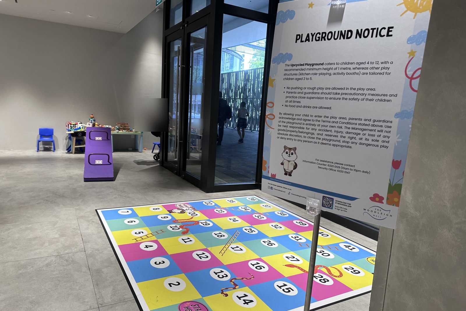 Playground entrance with the Playground Notice sign (rules + age guidance) and the Snakes & Ladders floor decal at the threshold. Through the glass door you can see the corridor to the rest of the mall.