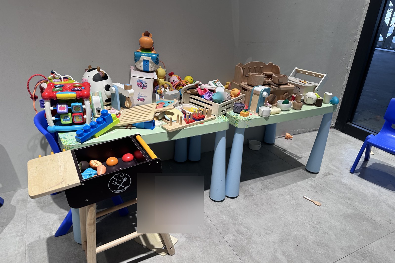 Toy table with wooden cooking tools, plastic learning toys, Fisher-Price-style activity cubes, a small wooden BBQ grill stool. Designed for the 2-5 age bracket.